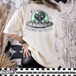 May include: A cream-coloured t-shirt featuring a graphic on the back of a grey alien inside a green-tinted UFO. The alien is making a peace sign with both hands. Below the UFO, the text reads I COME IN PEACE in a bold, dark grey font.