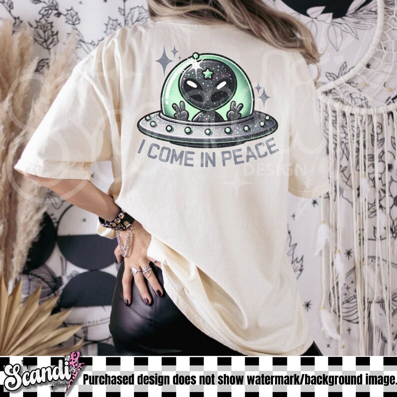 May include: A cream-coloured t-shirt featuring a graphic on the back of a grey alien inside a green-tinted UFO. The alien is making a peace sign with both hands. Below the UFO, the text reads I COME IN PEACE in a bold, dark grey font.