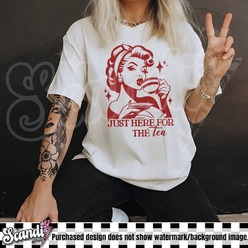 May include: Cream-colored t-shirt featuring a red graphic of a woman drinking tea, with the text "JUST HERE FOR THE TEA." The design has a retro aesthetic. The person is wearing black pants.