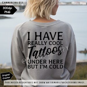May include: A heather grey long-sleeved sweatshirt featuring black text on the back that reads: I HAVE REALLY COOL Tattoos UNDER HERE BUT I'M COLD. The garment has a crew neckline and long sleeves, displaying a casual and functional design.