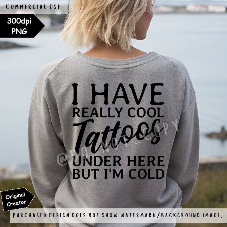 May include: A heather grey long-sleeved sweatshirt featuring black text on the back that reads: I HAVE REALLY COOL Tattoos UNDER HERE BUT I'M COLD. The garment has a crew neckline and long sleeves, displaying a casual and functional design.