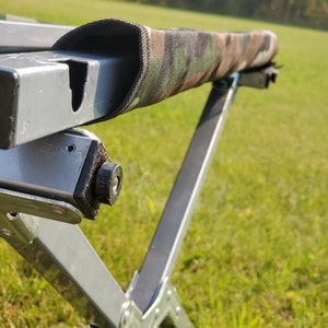 May include: A camouflage-patterned cover wrapped around a gray metal frame. The frame is part of a folding table or similar outdoor furniture, with a cross-legged design. The background shows a grassy field and trees.