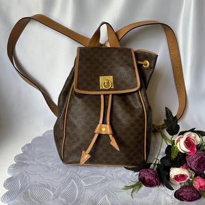 May include: Brown backpack with a repeating pattern, tan trim, and adjustable straps. The bag features a flap closure with a gold-tone clasp and drawstring closure. The backpack is set on a white lace doily with a bouquet of flowers.
