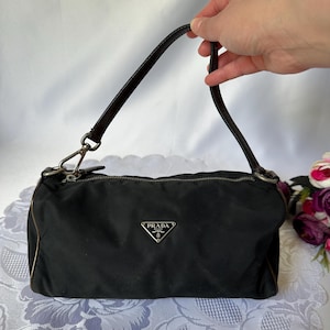 May include: Black Prada handbag with a silver-tone zipper and hardware. The bag has a leather handle and a small silver Prada logo on the front. The bag is rectangular and has a brown trim.