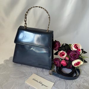 May include: A dark blue patent leather handbag with a silver-toned metal top handle featuring a repeating circular design. The bag includes a detachable shoulder strap and a white rectangular card with the text Salvatore Ferragamo printed on it.