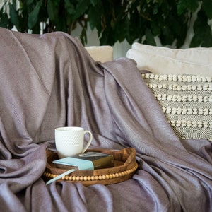 Könnte beinhalten: Eine weiche, lilafarbene Wolldecke, die über ein Sofa gelegt ist, mit einer weißen Tasse und einem Buch auf einem Holztablett. Das Sofa hat ein weißes Kissen und ein strukturiertes weißes Kissen mit schwarzen Akzenten.