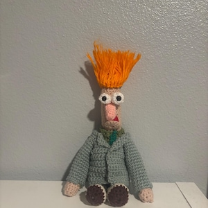 May include: A handmade crochet doll of Beaker from The Muppets. The doll has orange hair, large googly eyes, a pink nose, and a gray jacket. The doll is sitting on a white surface.