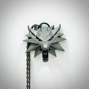 Puede incluir: Colgante plateado con la cabeza de un lobo con ojos rojos y un diseño geométrico. El colgante cuelga de una cadena oscura. La cabeza del lobo tiene la boca abierta y orejas puntiagudas.