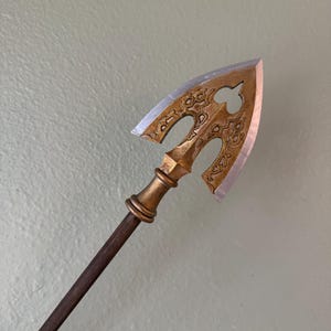 Puede incluir: Una flecha decorativa con una cabeza de color bronce y un asta marrón oscuro. La cabeza de la flecha presenta intrincados diseños recortados y una hoja afilada de color plateado. El asta es larga y delgada.