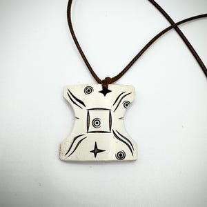 Op de afbeelding: Crèmekleurige hangerketting met een bruin koord. De hanger heeft een unieke vorm met zwart geëtste ontwerpen, waaronder cirkels, sterren en gebogen lijnen. De ketting wordt gepresenteerd tegen een witte achtergrond.