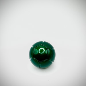 Puede incluir: Una esfera verde met&aacute;lico con un dise&ntilde;o geom&eacute;trico. La esfera tiene un acabado brillante y una serie de l&iacute;neas empotradas. La parte superior presenta un dise&ntilde;o hexagonal con una abertura circular. El objeto est&aacute; sobre un fondo blanco.