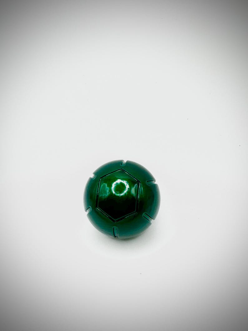 Puede incluir: Una esfera verde met&aacute;lico con un dise&ntilde;o geom&eacute;trico. La esfera tiene un acabado brillante y una serie de l&iacute;neas empotradas. La parte superior presenta un dise&ntilde;o hexagonal con una abertura circular. El objeto est&aacute; sobre un fondo blanco.