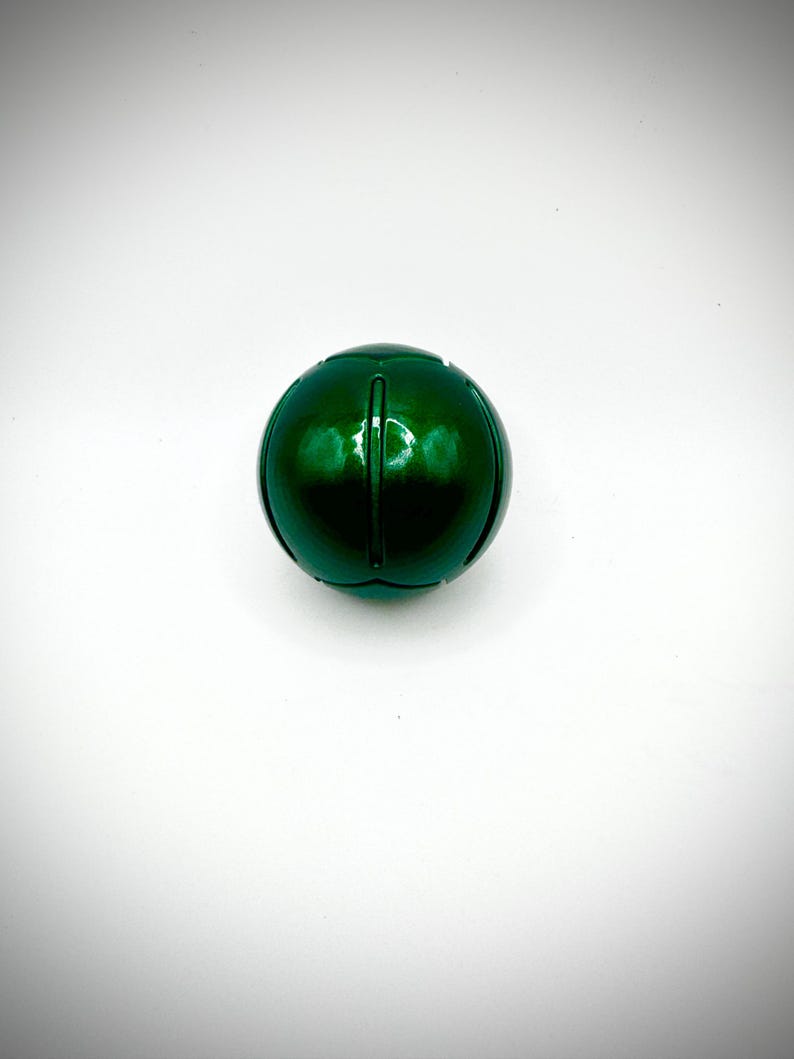 Puede incluir: Una esfera verde oscuro brillante con ranuras verticales. El objeto esf&eacute;rico est&aacute; centrado sobre un fondo blanco, mostrando su superficie reflectante y su forma redondeada. El objeto parece estar hecho de un material s&oacute;lido.