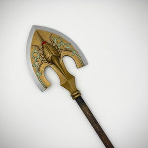May include: A prop arrow with a gold-colored arrowhead featuring a stylized design with teal accents and a red gem. The arrow has a wooden shaft with a dark brown finish. The arrowhead is approximately 6 inches wide.