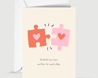 May include: Greeting card with a cream background and a matching envelope. The card features two puzzle pieces, one coral and one pink, each with a heart. The text reads, "Grateful we were written for each other."