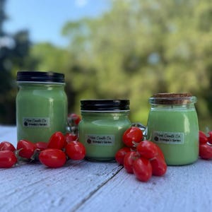 May include: Three green-colored candles in glass jars with black lids and a cork lid. The labels read "More Candle Co. Grandpa's Garden". Red cherry tomatoes are scattered around the candles on a white wooden surface.