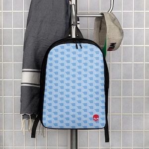 May include: A black backpack with a light blue and white geometric pattern. The backpack has black straps and a red skull logo on the lower right corner. The backpack is hanging on a metal rack.