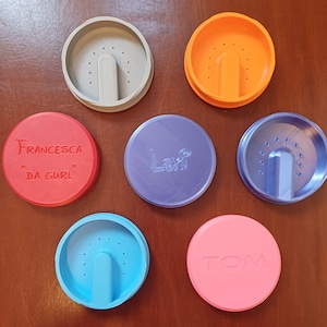 Pode incluir: Uma coleção de sete objetos circulares coloridos. Existem três recipientes abertos com uma protuberância semelhante a uma alça e uma série de pequenos orifícios. Quatro tampas sólidas também estão presentes, duas com os nomes "FRANCESCA DA GURL" e "TOM".