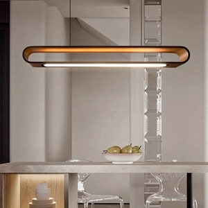 May include: A modern, rectangular wood and metal pendant light fixture with a warm white light source. The light is hanging over a table with clear chairs and a bowl of fruit.