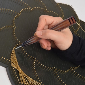 Könnte beinhalten: Ein dunkelgrünes String-Art-Stück aus Holz in einem floralen Design mit goldfarbenem Faden und kleinen Messingnägeln. Eine Person verwendet ein Holzwerkzeug, um den Goldfaden zu manipulieren und ein dekoratives Muster zu erstellen. Das Werkzeug hat einen dunklen Holzgriff.