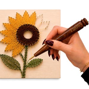 Könnte beinhalten: Ein quadratisches Holzbrett mit einem Sonnenblumen-Fadenkunst-Design, das aus gelbem, braunem und grünem Garn besteht, welches um Metallnägel gewickelt ist. Ein dunkelbraunes Holz-Punch-Needle-Werkzeug mit der Gravur Fältro ist abgebildet.