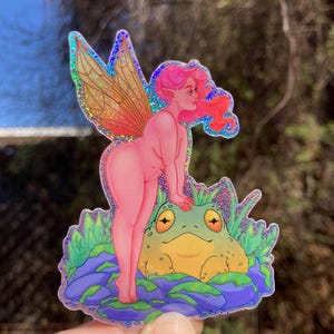 May include: A holographic sticker featuring a pink fairy with iridescent wings standing on a rock formation with a green frog. The fairy has pink hair and is looking to the right. The frog is sitting on the rock formation and has large, yellow eyes.