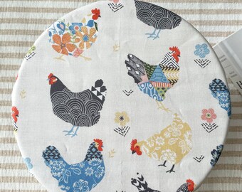 May include: Round white fabric bowl cover with a pattern of colorful chickens and flowers. The chickens are in various poses and have different patterns on their feathers. The flowers are in shades of pink, yellow, and orange.