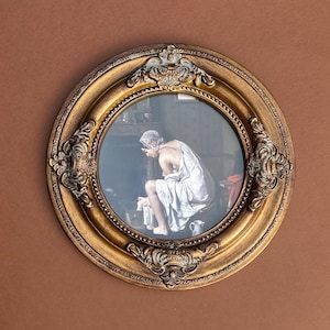 May include: A round, gold-colored framed print featuring a classical painting of a person in a white robe. The frame has ornate detailing and is set against a brown background. The painting depicts a person seated, with a jug nearby.