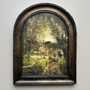 May include: A framed landscape painting showing a tranquil waterside scene. The artwork features a house, trees, and water, all in natural colours. The dark brown frame has an arched top, adding a classic touch to the artwork.