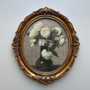 May include: An oval-shaped, gold-colored framed painting of white and pink roses in a dark green vase. The frame has ornate detailing and is set against a neutral background. The artwork evokes a classic, vintage aesthetic.