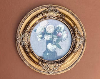 May include: A round, gold-colored framed print featuring a still life of white and pink roses in a dark blue vase. The ornate frame has detailed carvings and is set against a brown background.