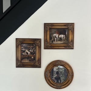 May include: Three framed artworks hang on a white wall. Two rectangular frames and one round frame with ornate, gold-toned detailing. The rectangular frames feature paintings, while the round frame is a mirror reflecting a person.