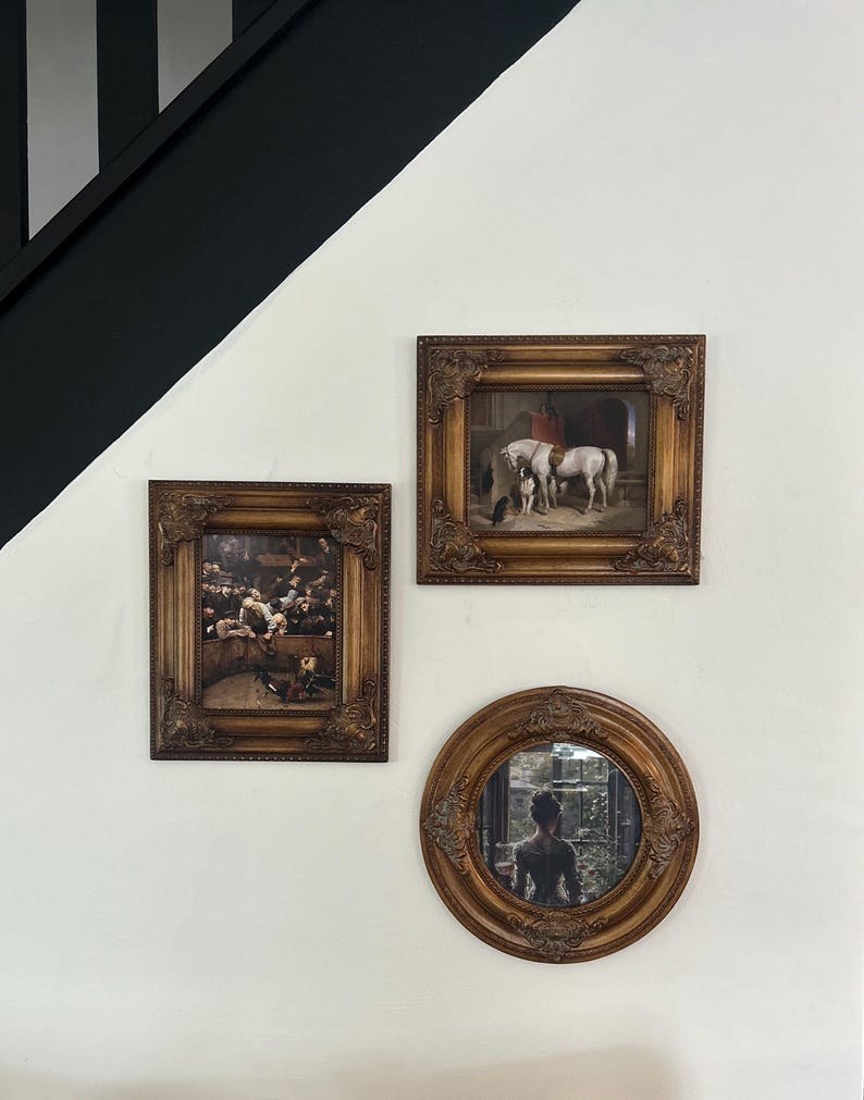 May include: Three framed artworks hang on a white wall. Two rectangular frames and one round frame with ornate, gold-toned detailing. The rectangular frames feature paintings, while the round frame is a mirror reflecting a person.