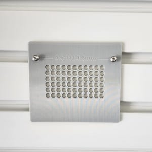 May include: A square, silver-colored metal vent cover with a grid of small, circular holes. The cover is secured with two screws and measures 4.5 inches (114.3mm) across. The background is white.