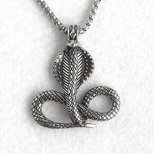 May include: A silver-toned metal necklace featuring a pendant shaped like a coiled cobra snake with a flared hood. The pendant is suspended from a thin, textured box chain. The metal has a dark, oxidized finish in the recessed areas to highlight the detailed scale texture of the snake's body and hood.