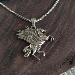 Könnte beinhalten: Halskette mit einem silberfarbenen Pegasus-Anhänger. Der Anhänger zeigt ein detailliertes geflügeltes Pferd. Die Halskette hat ein Kettenglied-Design. Das Wort "museum" steht auf einem kleinen schwarzen Etikett.