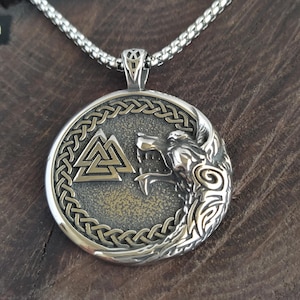 Può includere: Un ciondolo circolare color argento su una catenina abbinata, con un rilievo raffigurante una testa di lupo e un simbolo Valknut. Il ciondolo presenta uno sfondo metallico scuro e testurizzato, incorniciato da un bordo intrecciato.