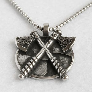 May include: A silver pendant necklace with a circular emblem and two crossed axes. The axes have detailed carvings on the blades and handles. The necklace chain is a delicate, linked design.