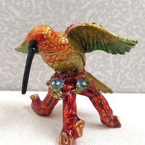 May include: A small, colourful hummingbird figurine with outstretched wings, perched on a red, twig-like base. The hummingbird is painted in shades of orange, yellow, and green, with black eyes and a long, black beak.