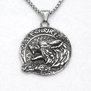 Könnte beinhalten: Eine silberfarbene Metall-Anhänger-Halskette mit einem kreisförmigen Design und einem detaillierten Wolfskopf im Profil. Das Wort FENRIR ist entlang des oberen Randes des Kreises eingraviert, der einen strukturierten Hintergrund und eine Box-Kette aufweist.