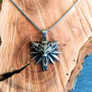 May include: A silver chain necklace with a wolf head pendant. The pendant is detailed with sharp teeth and a fierce expression. The wolf head is surrounded by a circle of spikes.