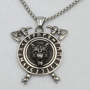 Puede incluir: Collar con colgante plateado con la cabeza de un lobo dentro de un círculo con runas y hachas cruzadas. El colgante cuelga de una cadena. Diseño de temática vikinga.