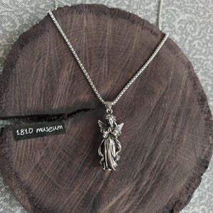 May include: A silver chain necklace with a pendant of a praying angel. The angel is wearing a long gown and has wings. The necklace is on a dark brown wooden surface. The text "1810 museum" is visible on a black tag.
