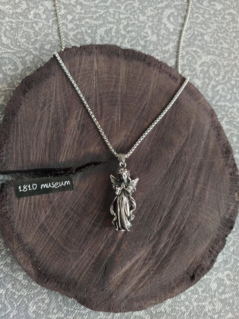 May include: A silver chain necklace with a pendant of a praying angel. The angel is wearing a long gown and has wings. The necklace is on a dark brown wooden surface. The text "1810 museum" is visible on a black tag.
