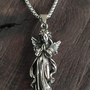 May include: A silver angel pendant necklace with a detailed design. The angel is depicted with wings, a flowing gown, and a crown of leaves. The necklace is made of a silver chain.