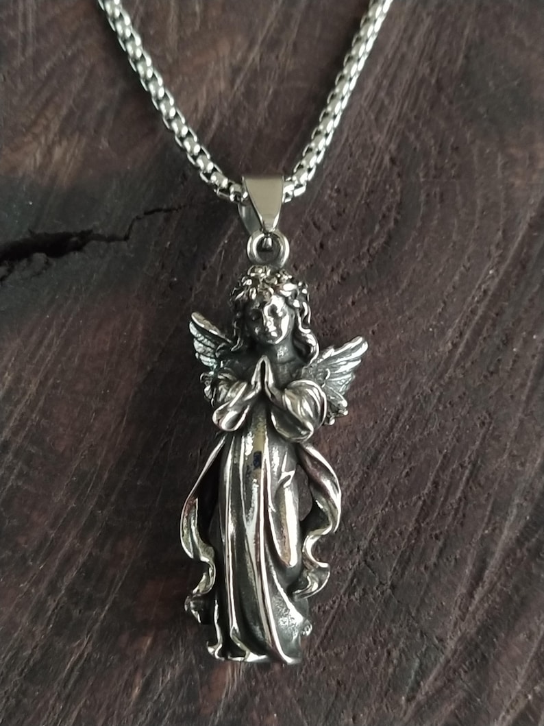May include: A silver angel pendant necklace with a detailed design. The angel is depicted with wings, a flowing gown, and a crown of leaves. The necklace is made of a silver chain.