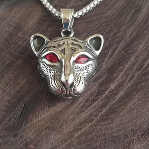 Puede incluir: Un collar con colgante plateado que presenta una detallada cabeza de leopardo con ojos de piedras preciosas rojas. El colgante está suspendido de una cadena plateada. El collar se exhibe sobre una superficie de madera oscura.