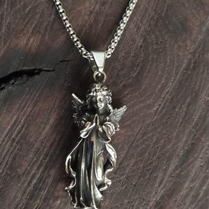May include: A silver chain necklace with a detailed silver angel pendant. The angel is depicted with wings, a flowing gown, and hands clasped in prayer.