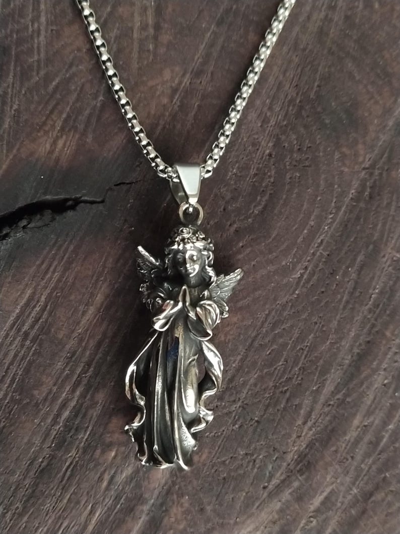 May include: A silver chain necklace with a detailed silver angel pendant. The angel is depicted with wings, a flowing gown, and hands clasped in prayer.