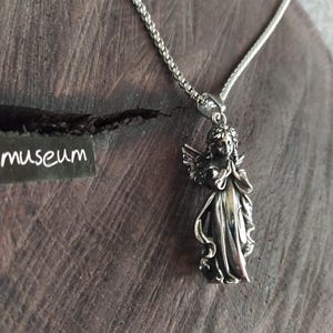 May include: A silver chain necklace with a pendant of a silver angel with wings outstretched. The angel is wearing a flowing robe and has a serene expression on its face.